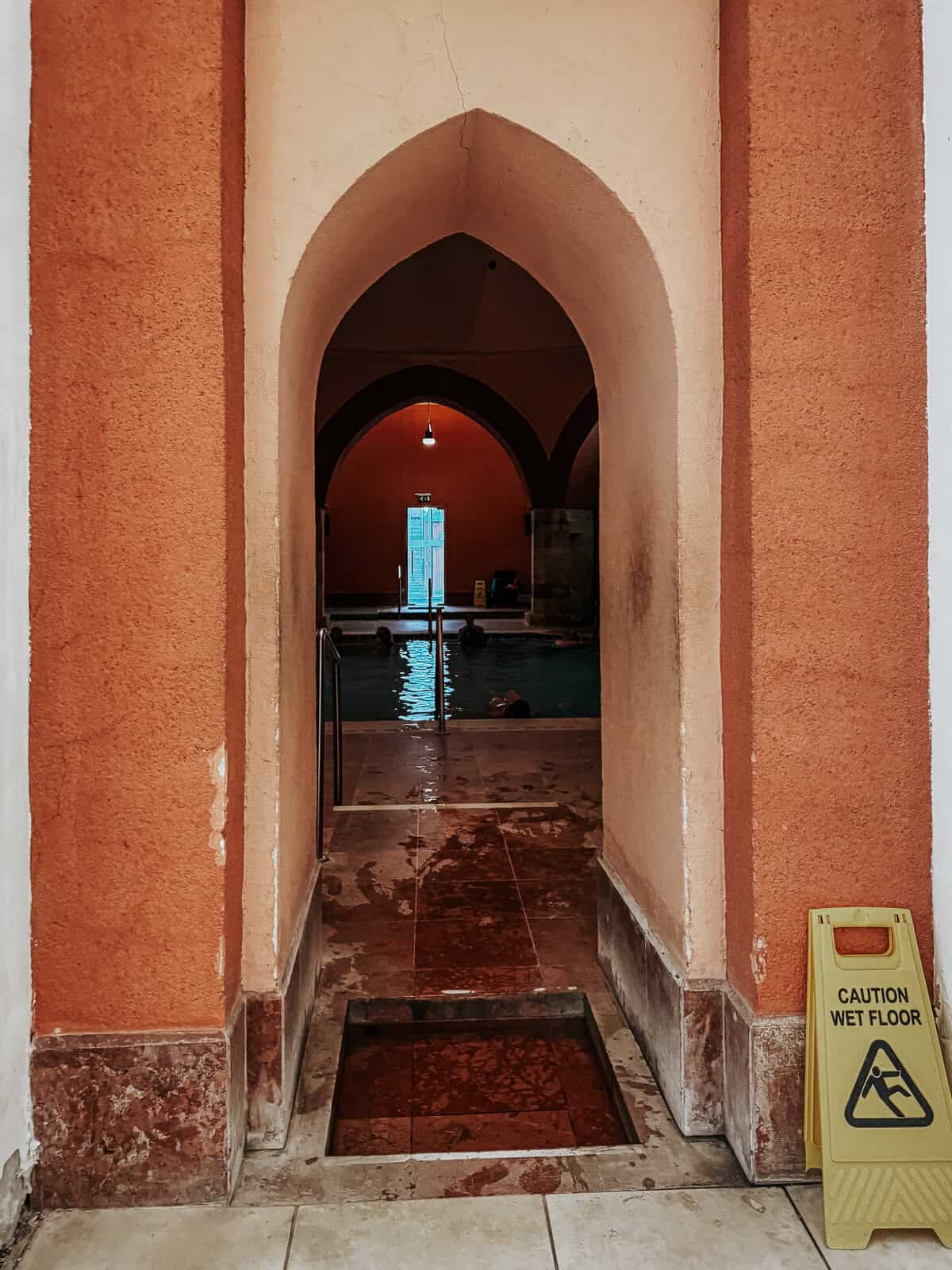 A view through an arched doorway into a swimming pool area, with a "Caution Wet Floor" sign placed near the entrance and some people swimming in the pool.