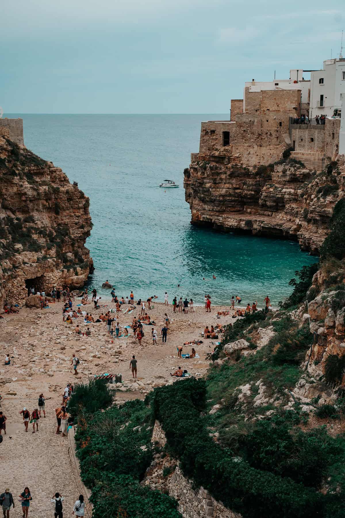 A crowded pebble beach nestled between dramatic cliffs in Polignano a Mare, Italy. People are sunbathing, swimming, and enjoying the scenic blue waters. The historic buildings perched on the cliffside add to the charm of this famous coastal town