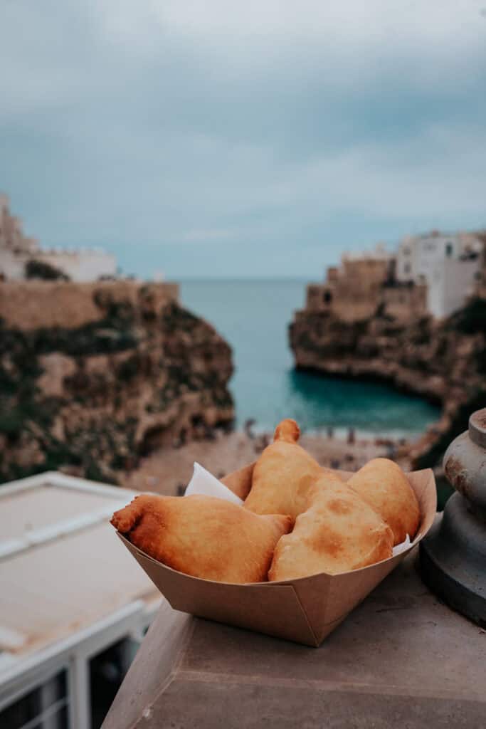 A close-up of a paper bowl filled with golden, crispy panzerotti (fried stuffed pastries), placed on a stone ledge. In the blurred background, the iconic cliffs and turquoise waters of Polignano a Mare, Italy, create a picturesque setting.
