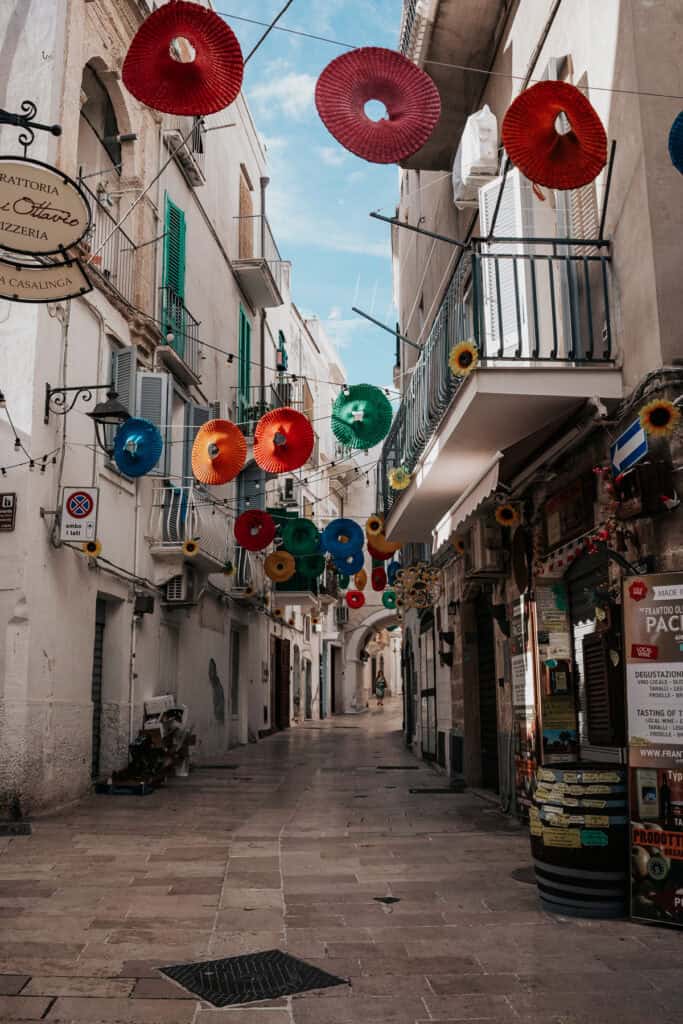 A charming narrow street in Monopoli, Italy, decorated with colorful paper lanterns in red, blue, green, and yellow, strung overhead. Traditional whitewashed buildings with green shutters and small balconies line the street, while local shops and trattorias add to the cozy, inviting atmosphere. A lone pedestrian walks towards the end of the alley, enhancing the scene’s picturesque charm