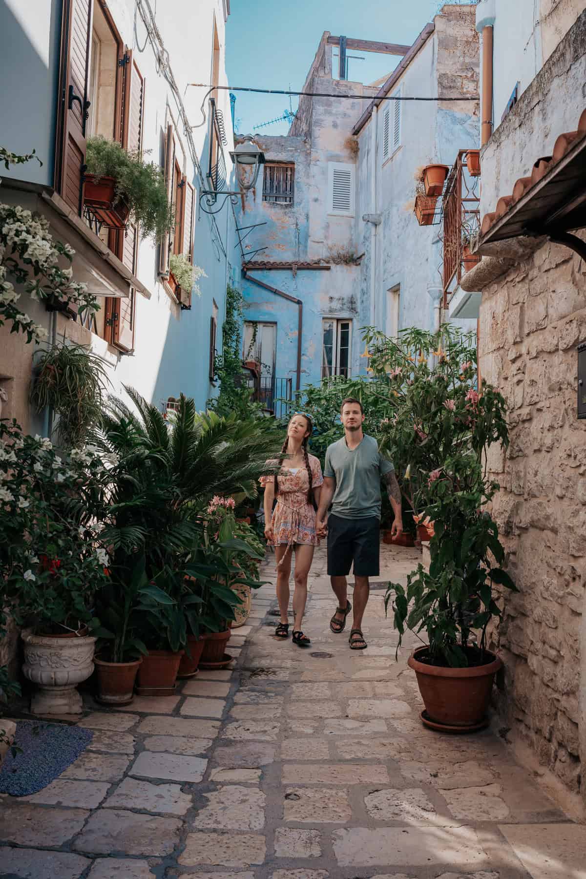 A young couple holding hands while strolling through a narrow alley in Casamassima, Italy, surrounded by lush potted plants. The pastel-colored walls, rustic shutters, and a mix of stone and plaster buildings create a romantic, old-world charm.