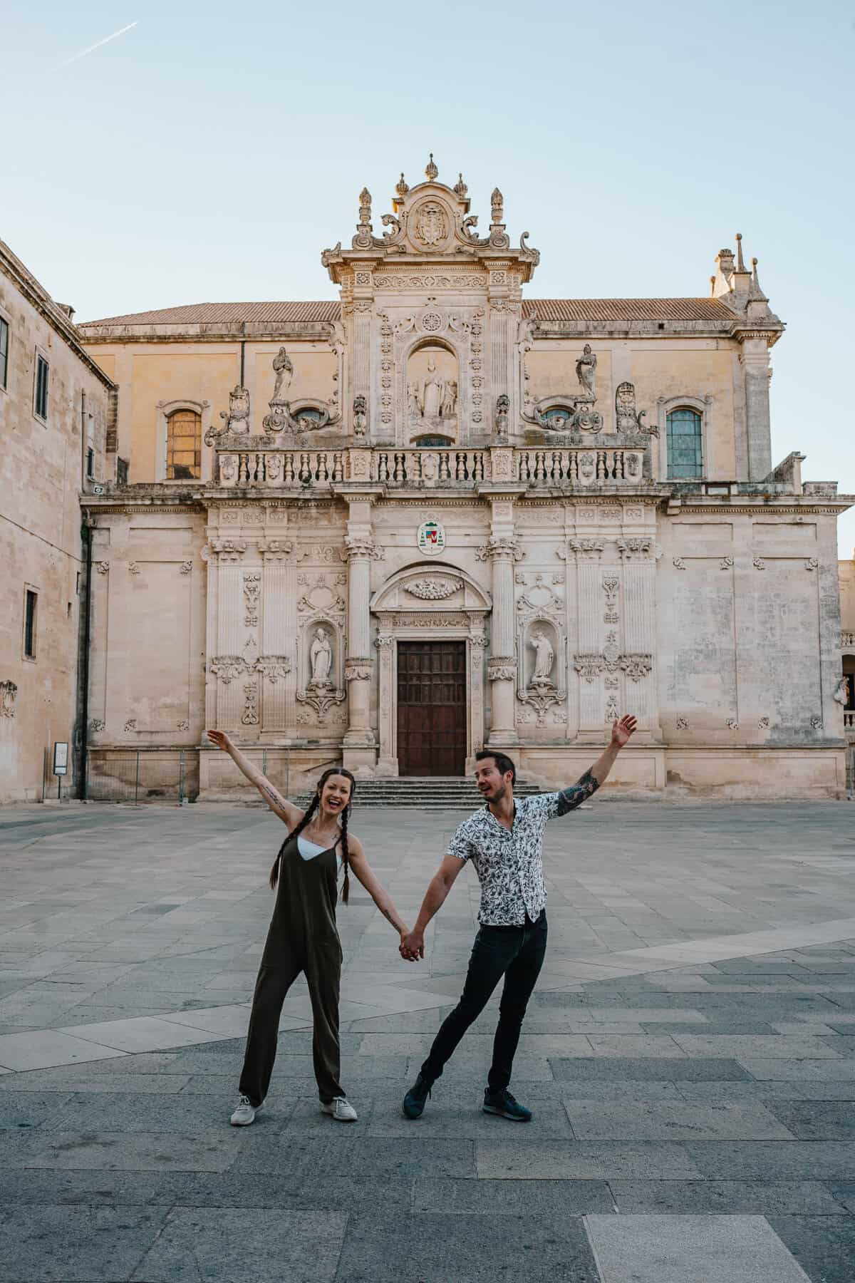 A cheerful couple holding hands and playfully dancing in front of the ornate baroque façade of a historic church in Lecce, Italy. The grand architecture, complete with intricate carvings and statues, contrasts with their joyful energy.