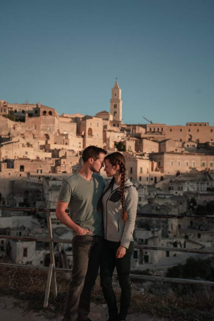 A young couple stands close together at sunset in Matera, Italy, gazing into each other's eyes. The ancient cityscape of stone houses and the bell tower of Matera Cathedral rise behind them, bathed in golden light.