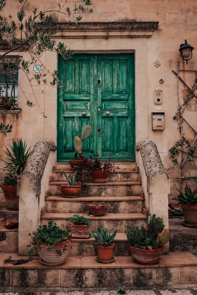 A beautifully weathered green wooden door sits atop a small stone staircase in Matera, Italy, surrounded by potted plants and succulents. The aged limestone walls and rustic charm create an inviting Mediterranean feel.