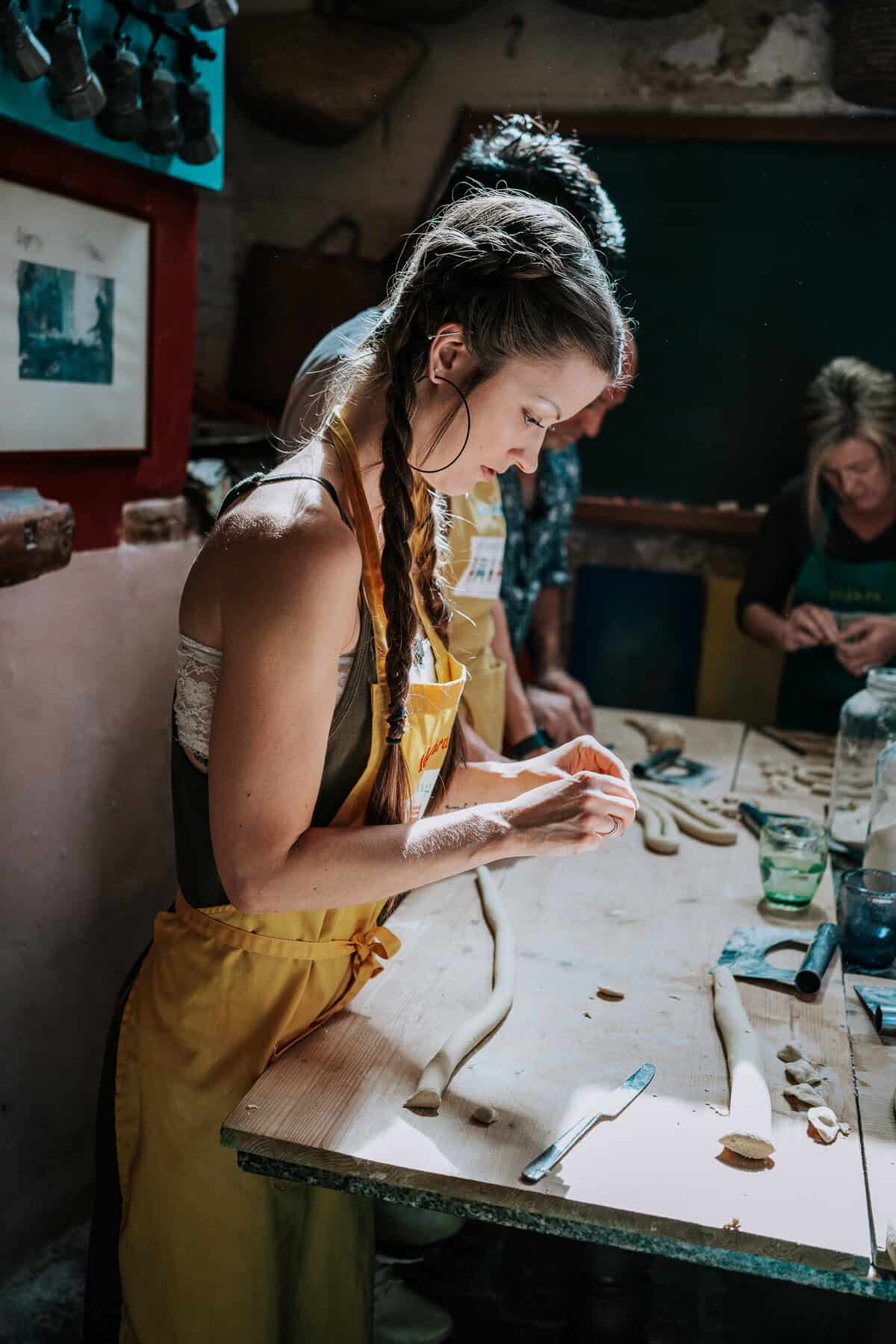 A woman wearing a yellow apron and with long braided hair focuses on shaping fresh pasta at a wooden table. The warm lighting and rustic kitchen setting enhance the traditional Italian culinary experience.