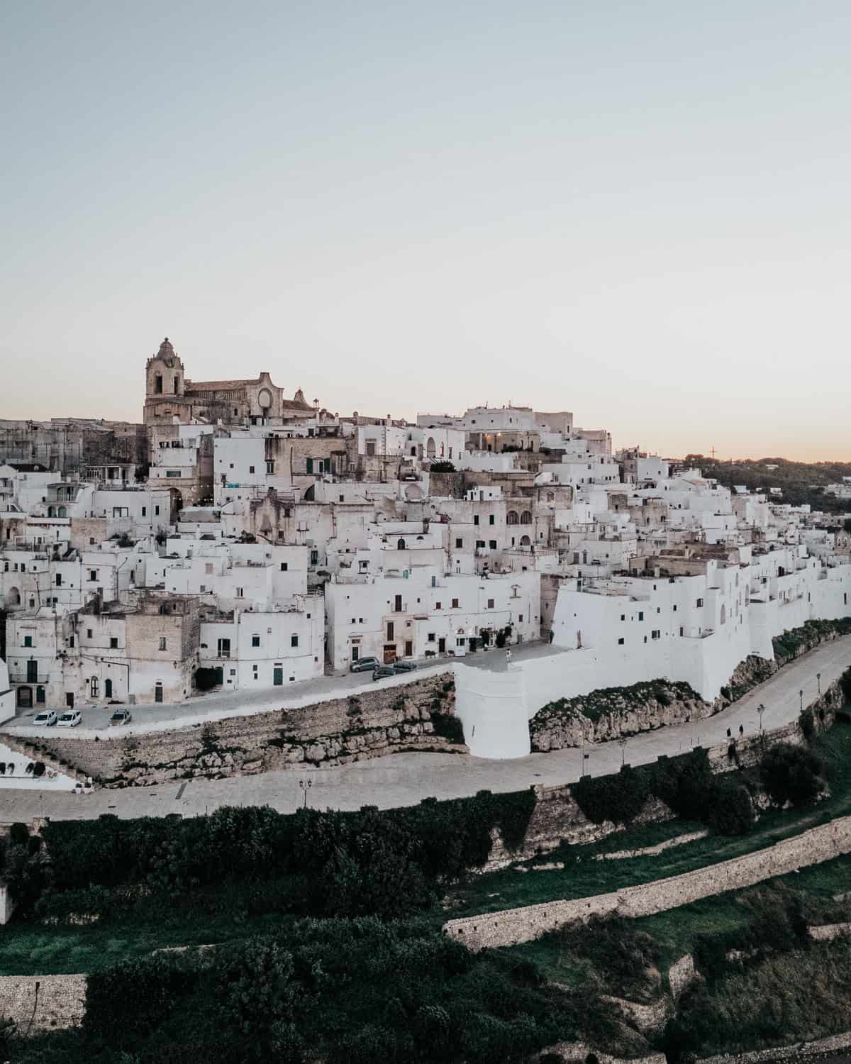 A breathtaking aerial view of Ostuni, Italy, also known as the "White City," with its iconic whitewashed buildings cascading across the hillside. A historic church stands at the highest point, overlooking winding streets and lush greenery.