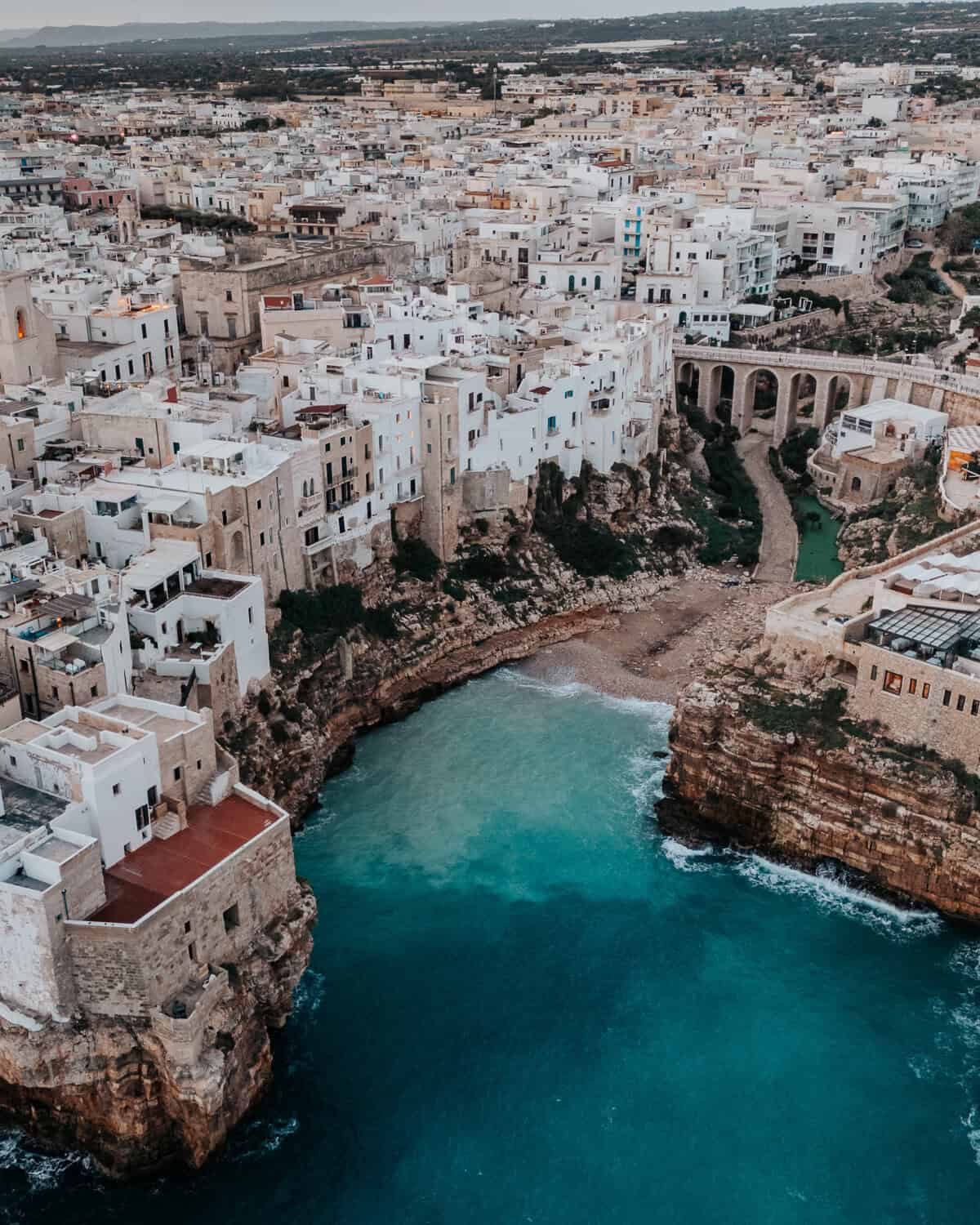 A breathtaking aerial capture of Polignano a Mare, a cliffside town in Italy, with turquoise waters crashing against rugged limestone cliffs and traditional white buildings.