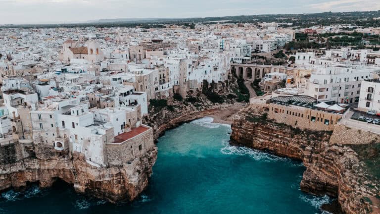 A breathtaking aerial capture of Polignano a Mare, a cliffside town in Italy, with turquoise waters crashing against rugged limestone cliffs and traditional white buildings.