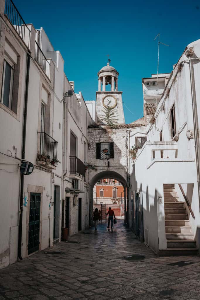 A narrow cobblestone alley in Casamassima, Italy, framed by historic white buildings, with a clock tower visible in the background