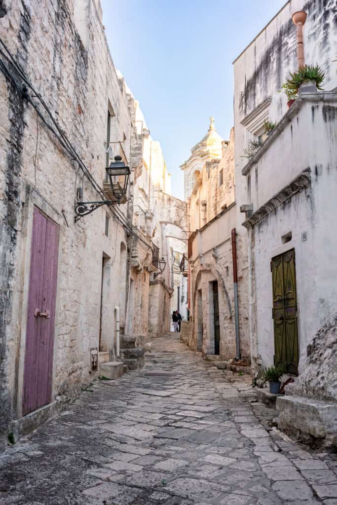A historic stone-paved street in Ceglie Messapica, Italy, flanked by ancient limestone buildings with vibrant wooden doors.