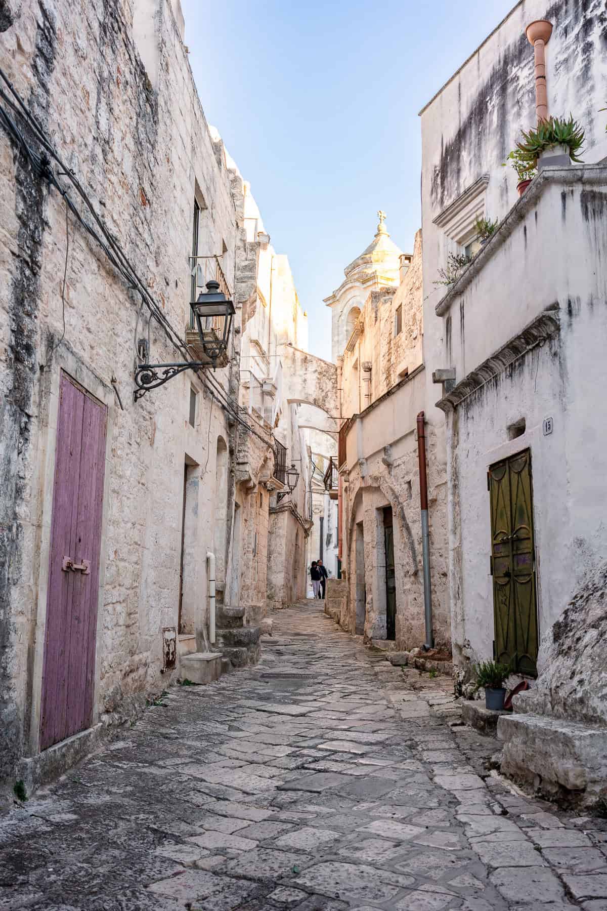 A historic stone-paved street in Ceglie Messapica, Italy, flanked by ancient limestone buildings with vibrant wooden doors.