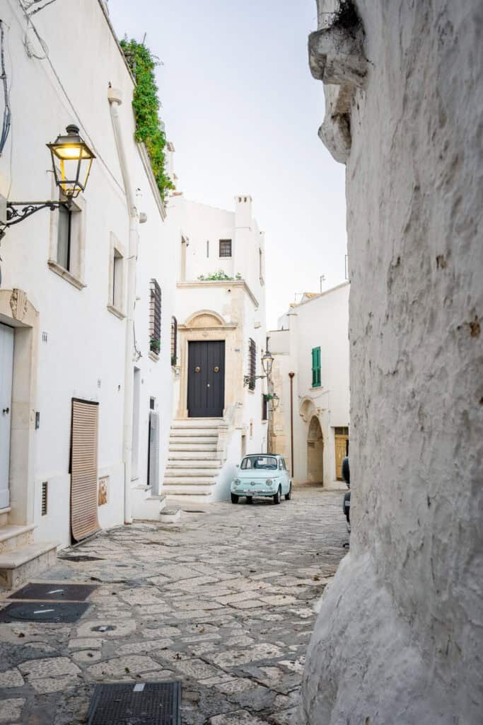 A scenic view of an old town street with winding stone pathways, traditional architecture, and a vintage car parked nearby.