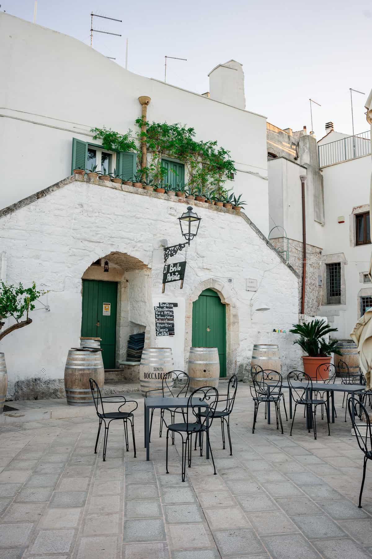 A small outdoor café in a historic whitewashed building with green doors and wooden barrels, exuding a rustic Mediterranean charm.