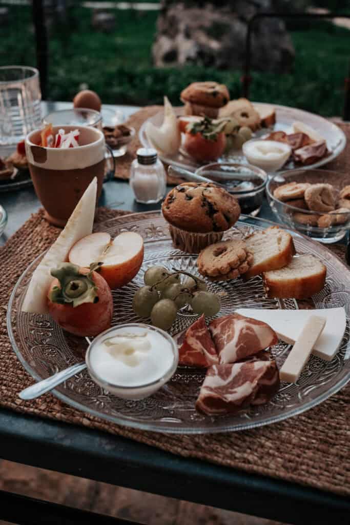 A breakfast spread in a beautiful garden featuring yogurt, a muffin, some cookies, cheese nad ham