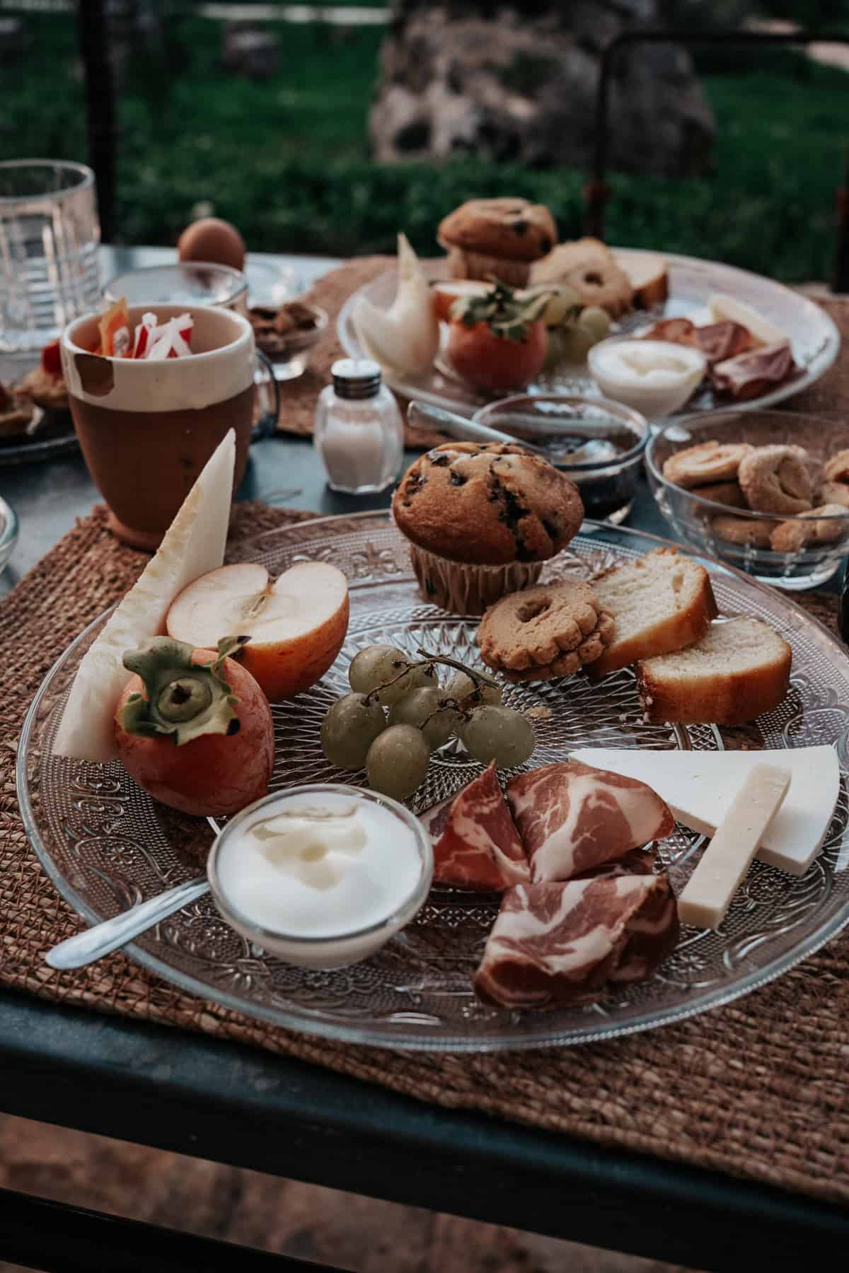 A breakfast spread in a beautiful garden featuring yogurt, a muffin, some cookies, cheese nad ham
