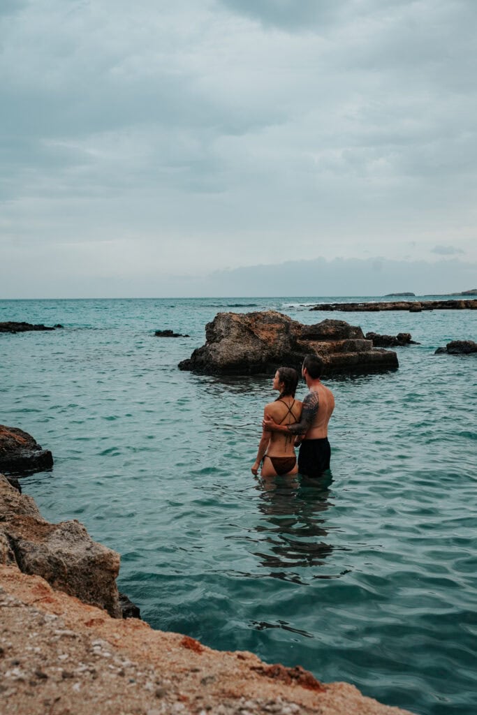 A couple stands in the shallow waters of Polignano a Mare, admiring the view of the Adriatic under an overcast sky.