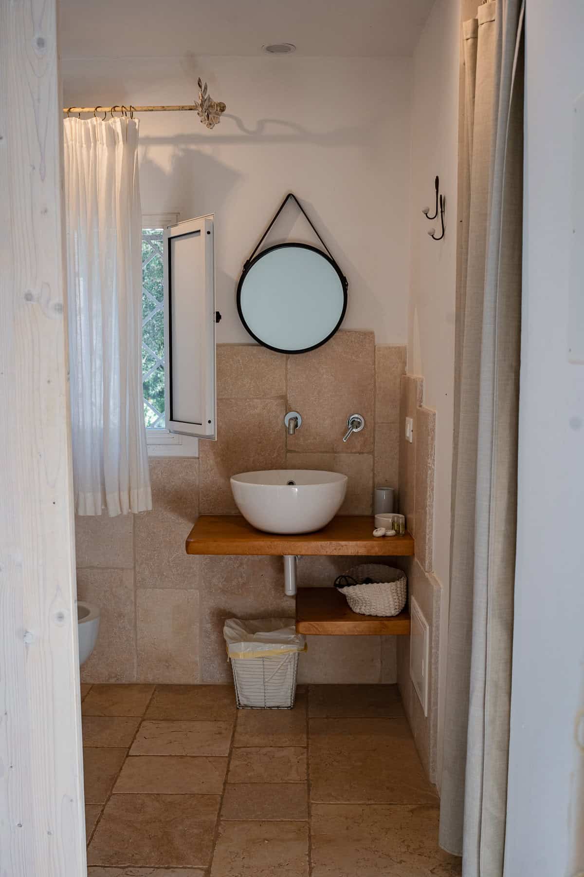 A stylish hotel bathroom with beige stone tiles, a white bowl sink on a wooden counter, and a round hanging mirror.