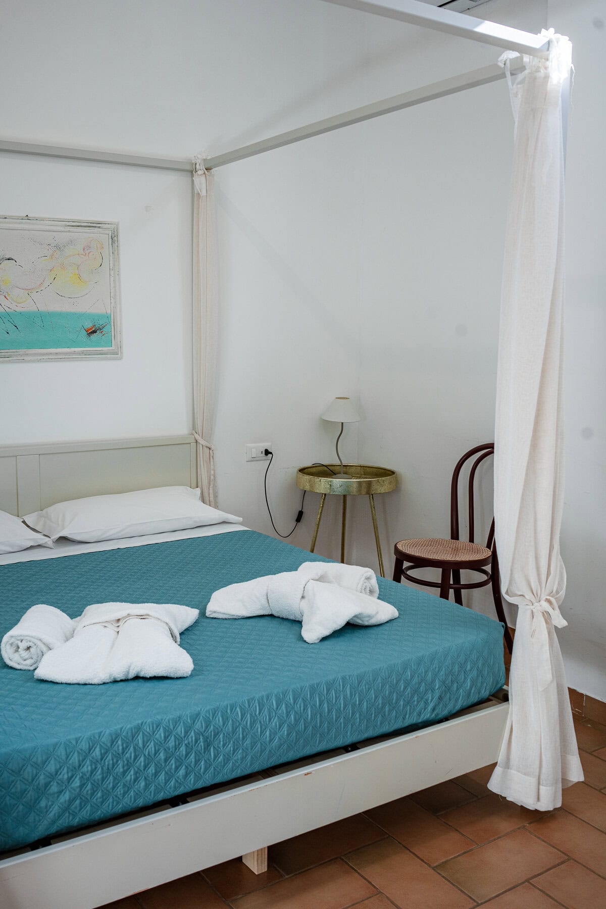 A minimalist hotel bedroom with a four-poster bed, teal bedding, and towels folded into decorative shapes.