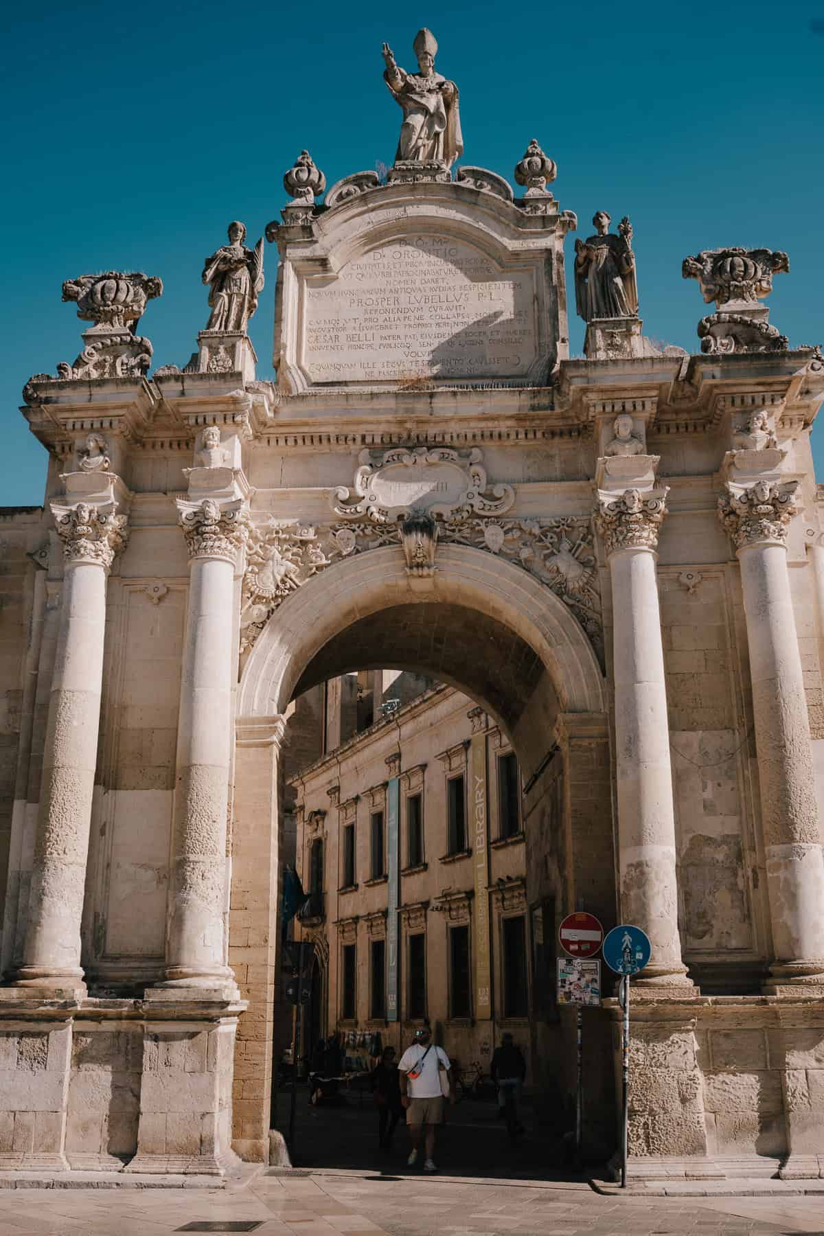 The grand Porta Napoli in Lecce, an ornate triumphal arch with detailed carvings and statues, standing under a bright blue sky.