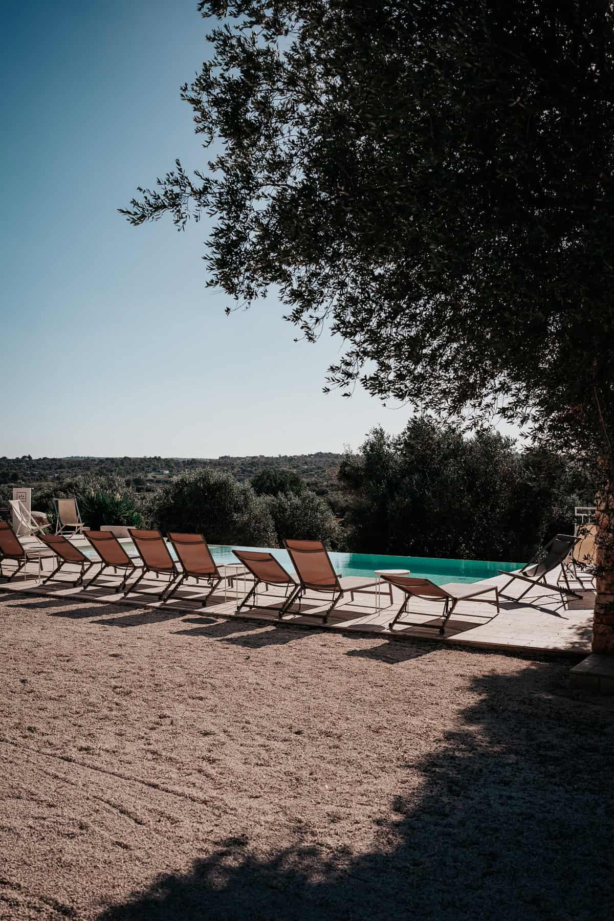 An idyllic infinity pool in Ostuni, Italy, surrounded by sun loungers and lush olive trees, offering a serene countryside escape.