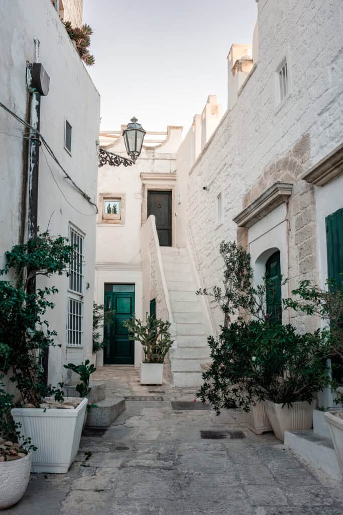 A classic whitewashed building in Ostuni, Italy, with vibrant green doors and blooming pink flowers cascading over the steps.