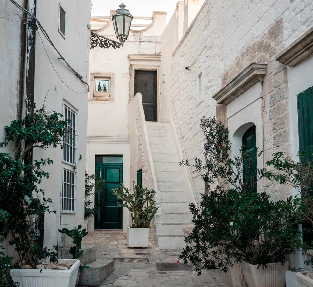 A classic whitewashed building in Ostuni, Italy, with vibrant green doors and blooming pink flowers cascading over the steps.