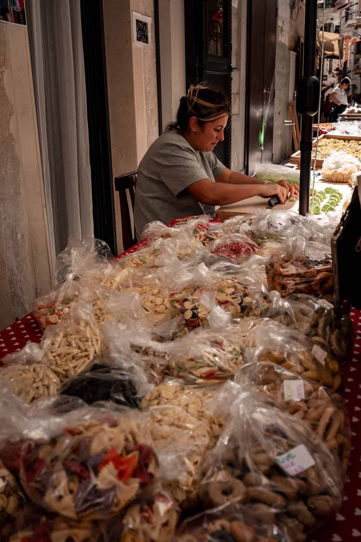 A woman in a grey shirt sits at a table in an outdoor market, skillfully making fresh pasta by hand. In the foreground, a table covered with a red polka-dot cloth is filled with plastic bags of various dried pastas and baked goods. The scene is set in a narrow Italian street with more market stalls in the background.