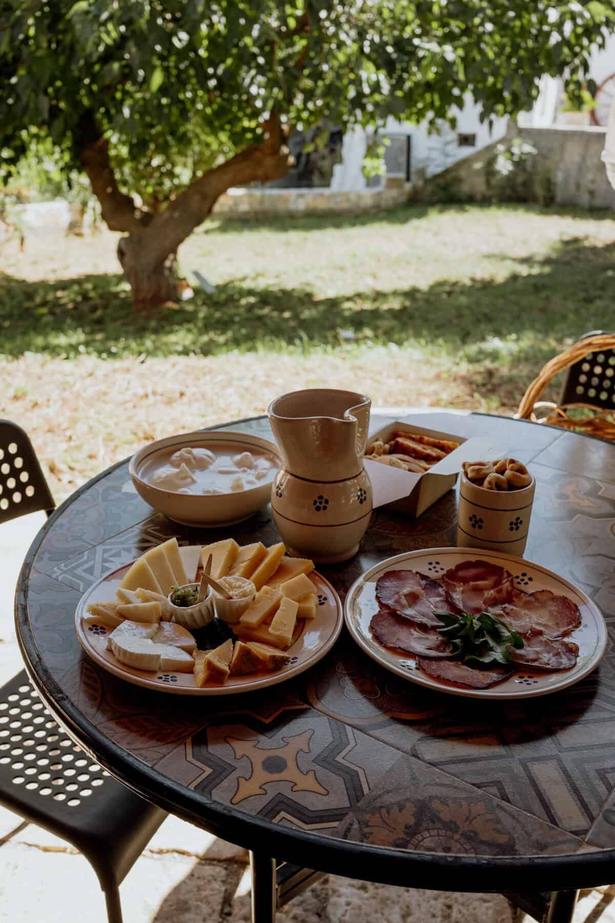 A rustic outdoor table set under the shade of a tree, featuring a spread of local cheeses, cured meats, and a ceramic pitcher of wine. The cheese plate includes various types of sliced and cubed cheeses, served with a small bowl of condiments. Another plate holds thinly sliced cured meats garnished with fresh greens, while small bowls of snacks and a box of pastries complete the inviting countryside meal.