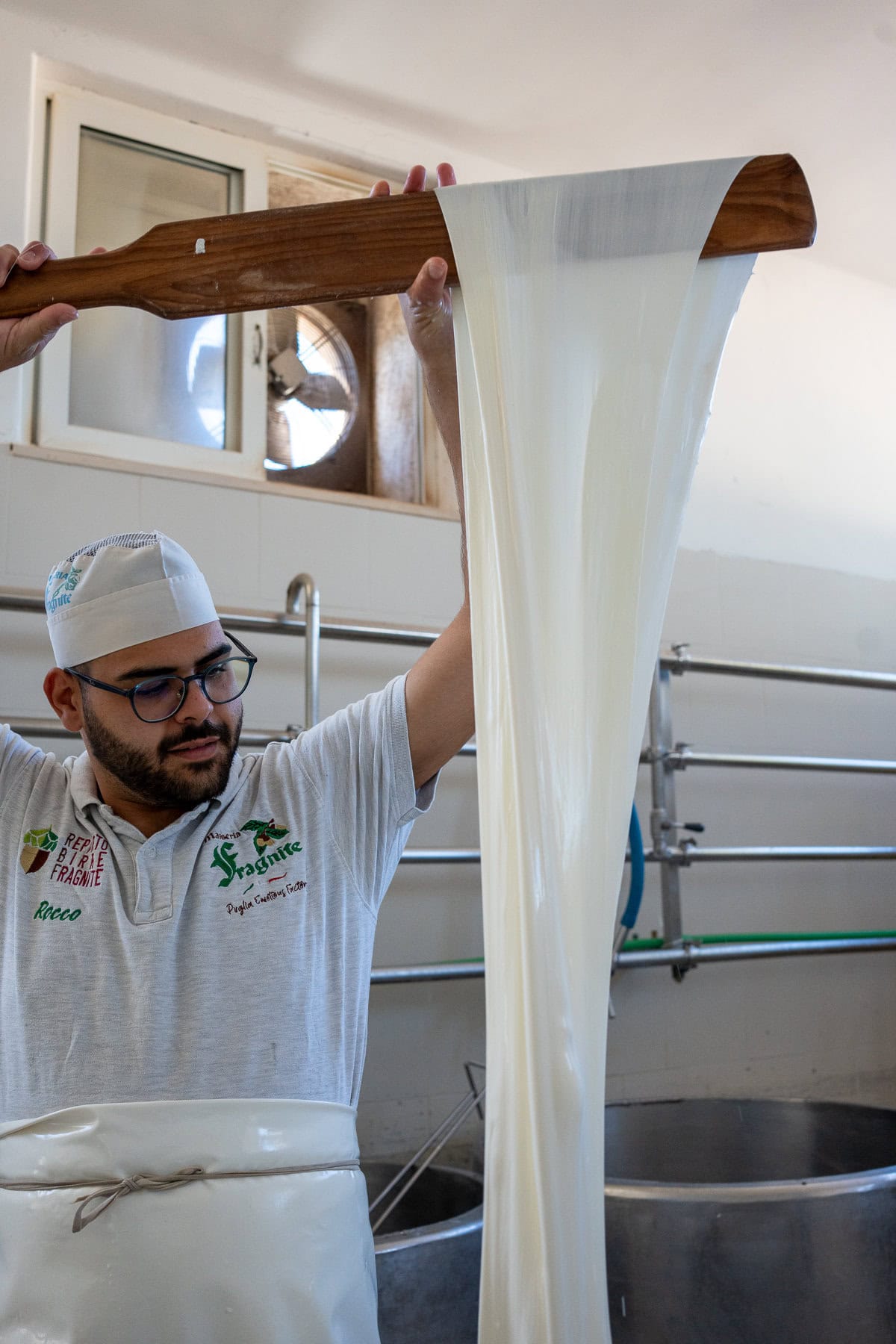 A cheesemaker wearing glasses, a white apron, and a branded polo shirt lifts a long, silky stretch of fresh mozzarella curd with a wooden paddle. The process takes place in a dairy facility with stainless steel equipment and a ventilation fan in the background.