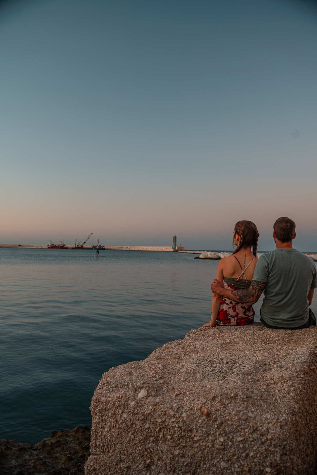 A couple sits on a rocky ledge overlooking the calm sea at sunset. The woman wears a floral dress, and the man, with a tattooed arm, embraces her as they watch the horizon together. A pier with a lighthouse and construction cranes stretches into the water in the background.