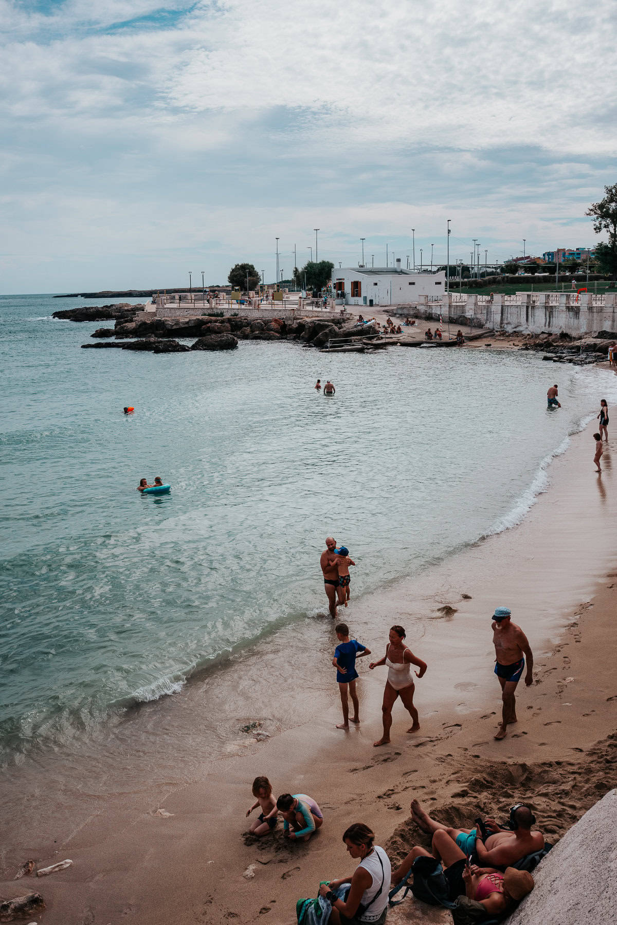 A sandy beach along the turquoise coastline, where families and swimmers enjoy the shallow waters. People of all ages wade in the sea, lounge on the shore, and play in the sand, while a stone jetty and small white buildings line the background.