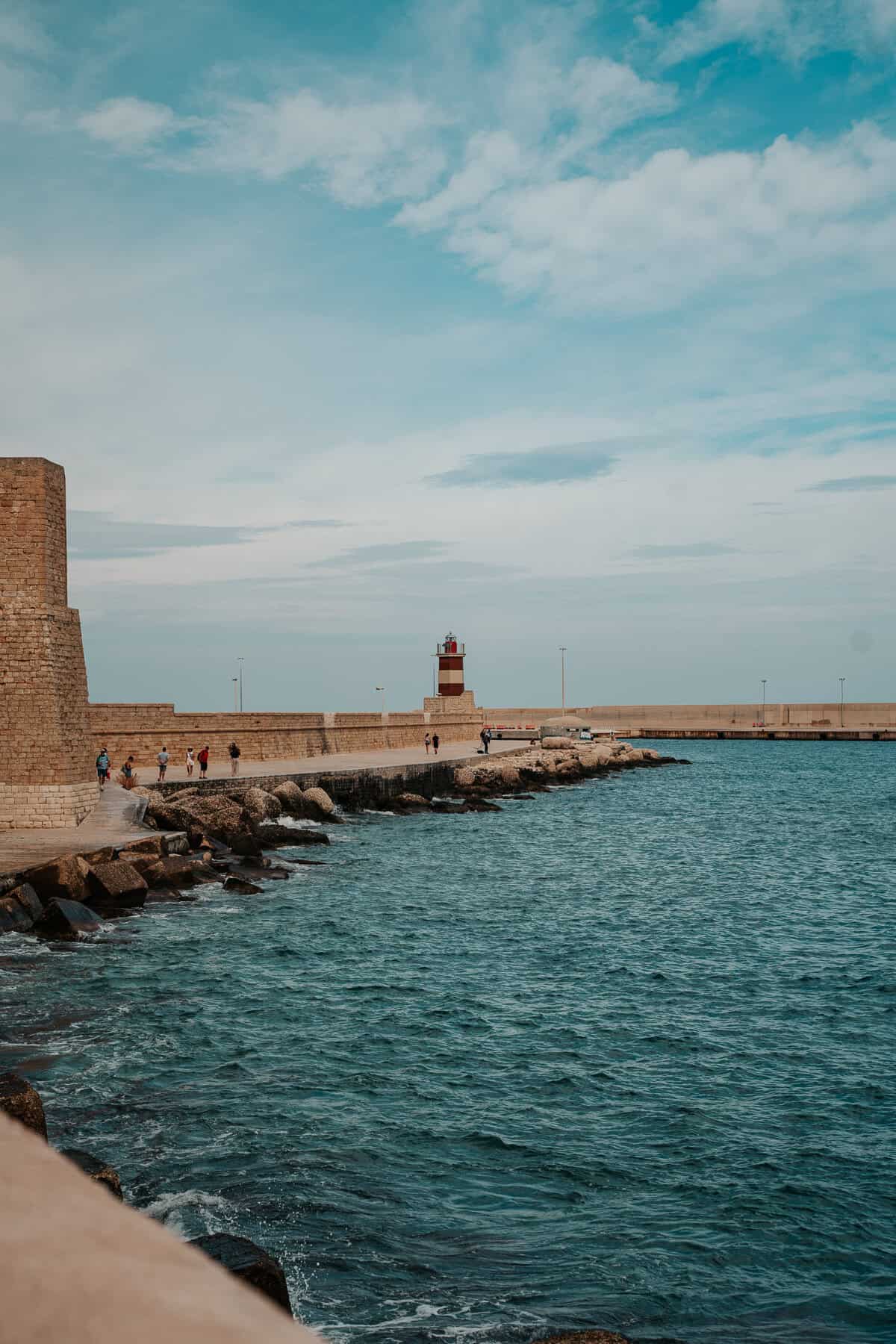 A scenic seaside pier with a red and white lighthouse standing at the end of a stone breakwater. The blue waters of the sea gently lap against the rocky shoreline, while a few people stroll along the path, enjoying the coastal view.