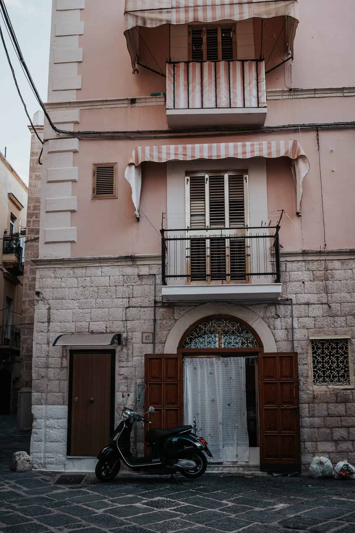 A charming Italian building with a pink facade, featuring striped awnings over its balconies and windows. A black Vespa scooter is parked in front of the wooden double doors, while a few bags of trash sit on the stone pavement nearby.