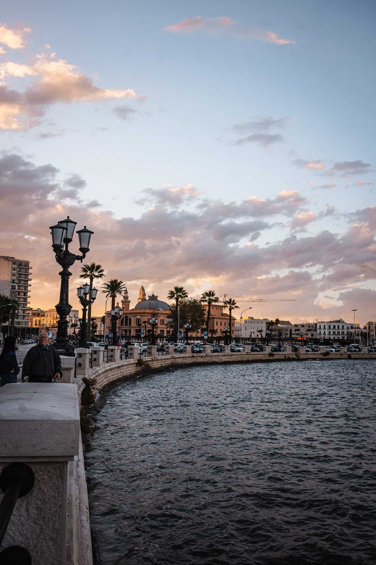A waterfront promenade lined with lampposts, palm trees, and historic buildings during a golden sunset. The rippling water reflects the warm hues of the sky, while people stroll along the railing, enjoying the view.