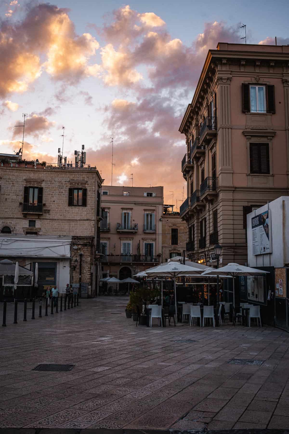 A lively European square at sunset, surrounded by historic buildings with elegant architecture. Outdoor café tables with white umbrellas sit in the center, while a few people walk through the cobblestone plaza under a sky painted with golden clouds.