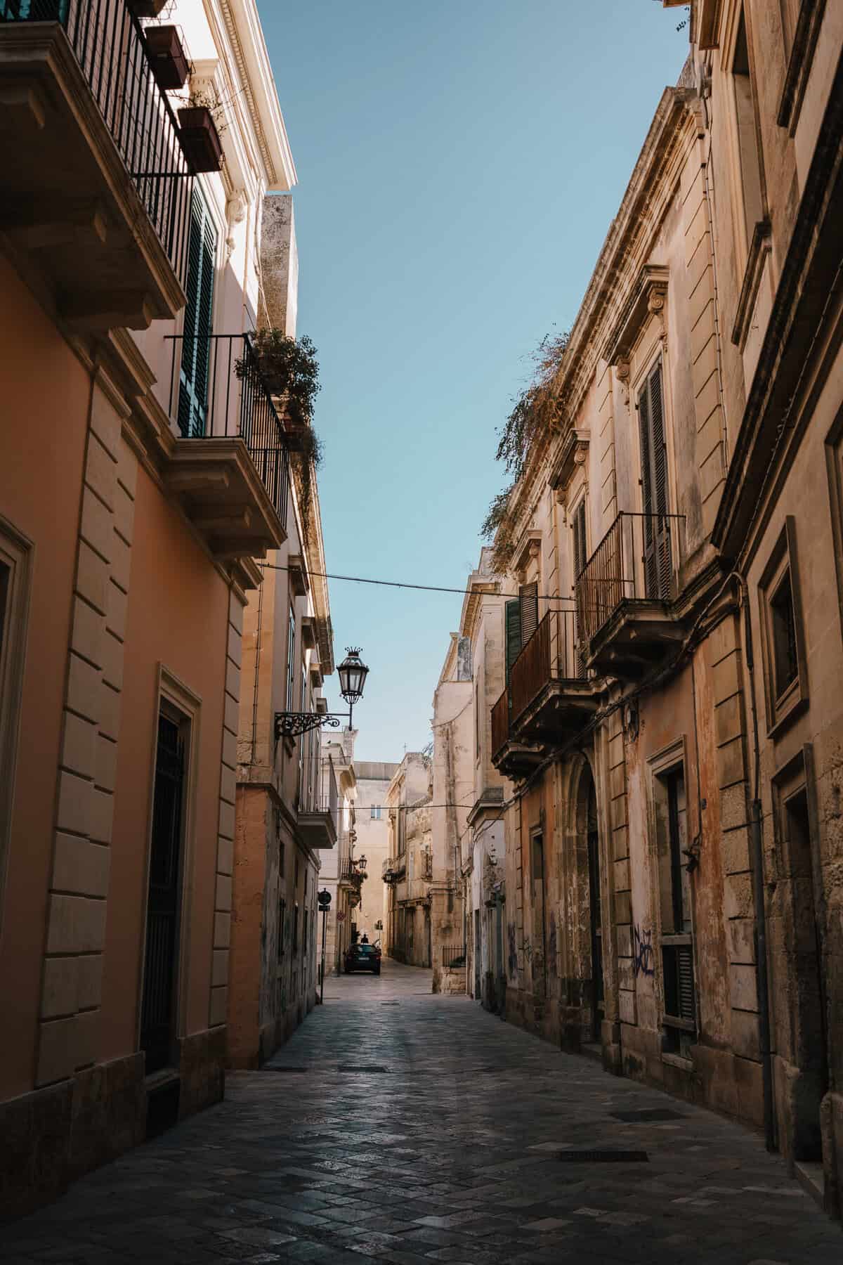 A quiet, narrow alleyway in a historic Italian town, lined with aged buildings adorned with wrought iron balconies and potted plants. The soft sunlight highlights the warm hues of the stone facades, leading the eye toward the vanishing point where a small car is parked.