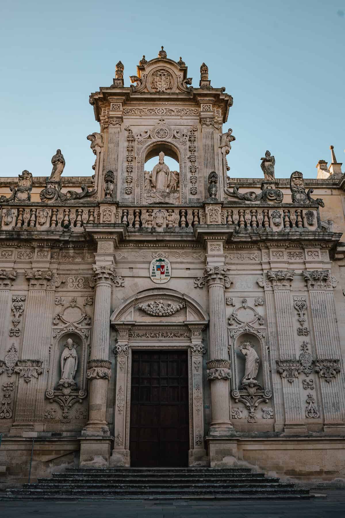 The ornate façade of a grand Baroque-style church, adorned with intricate carvings, statues, and religious iconography. A large wooden door sits at the entrance, with a row of sculpted figures and decorative elements embellishing the upper level.