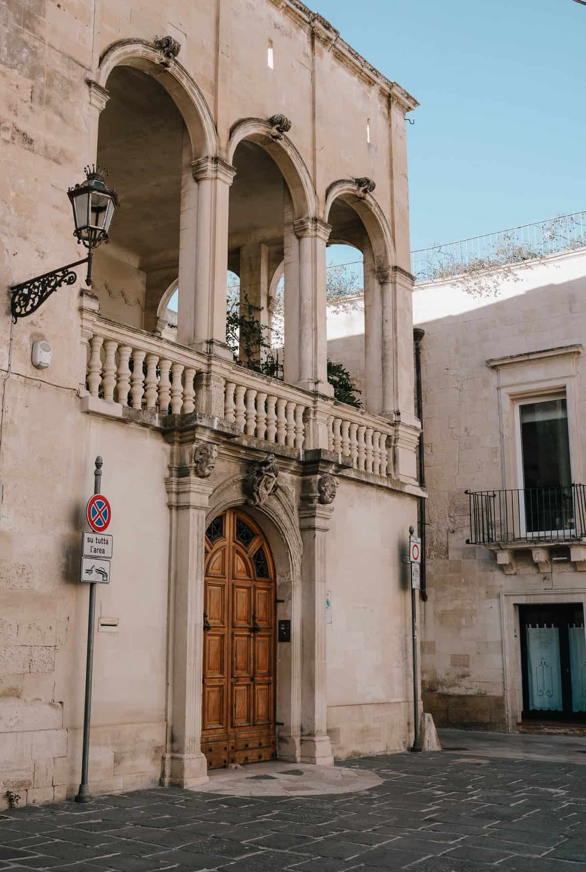 A historic building with grand arched openings and intricate stone detailing, showcasing classic Italian architecture. A wooden door beneath the arches serves as the main entrance, while a "no parking" sign stands nearby on the cobblestone street.