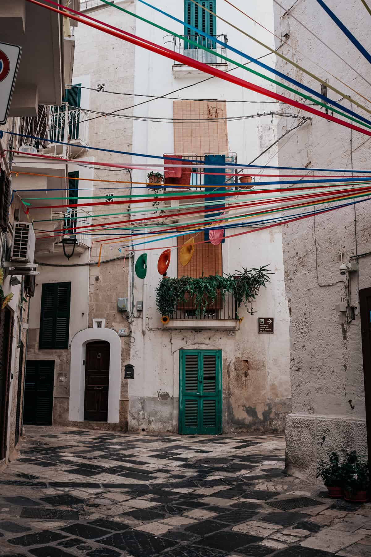 A narrow cobblestone street in a Mediterranean town, lined with whitewashed buildings adorned with sunflowers and colorful paper decorations. Vibrant red, blue, green, and yellow paper lanterns hang above, creating a festive atmosphere under a bright blue sky.