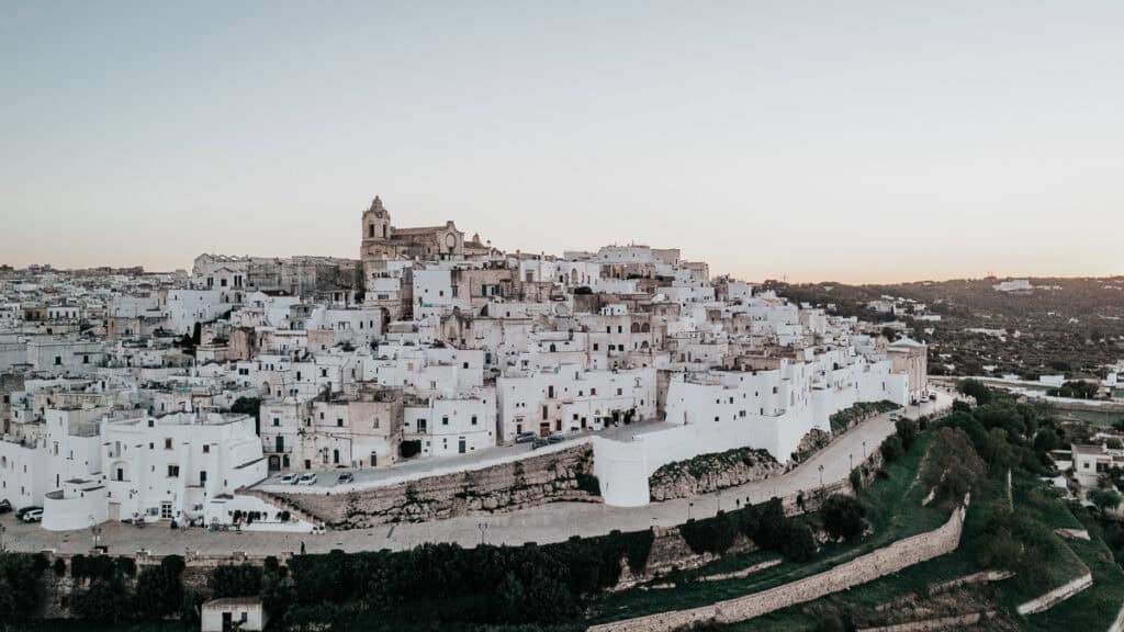 A breathtaking view of Ostuni, the whitewashed city in Puglia, perched on a hill at sunset. The densely packed historic buildings glow in the fading light, overlooking the lush green landscape below.