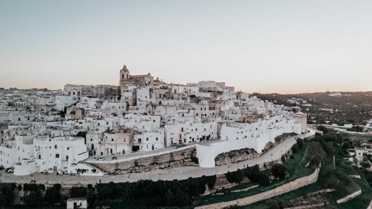 A breathtaking view of Ostuni, the whitewashed city in Puglia, perched on a hill at sunset. The densely packed historic buildings glow in the fading light, overlooking the lush green landscape below.