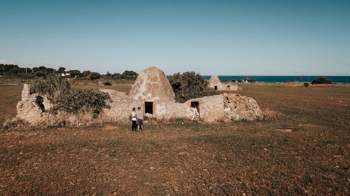 An old stone structure with conical roofs, possibly traditional trulli or rural farm buildings, stands in an open field near the sea. A few people explore the ruins, with the blue horizon in the distance.