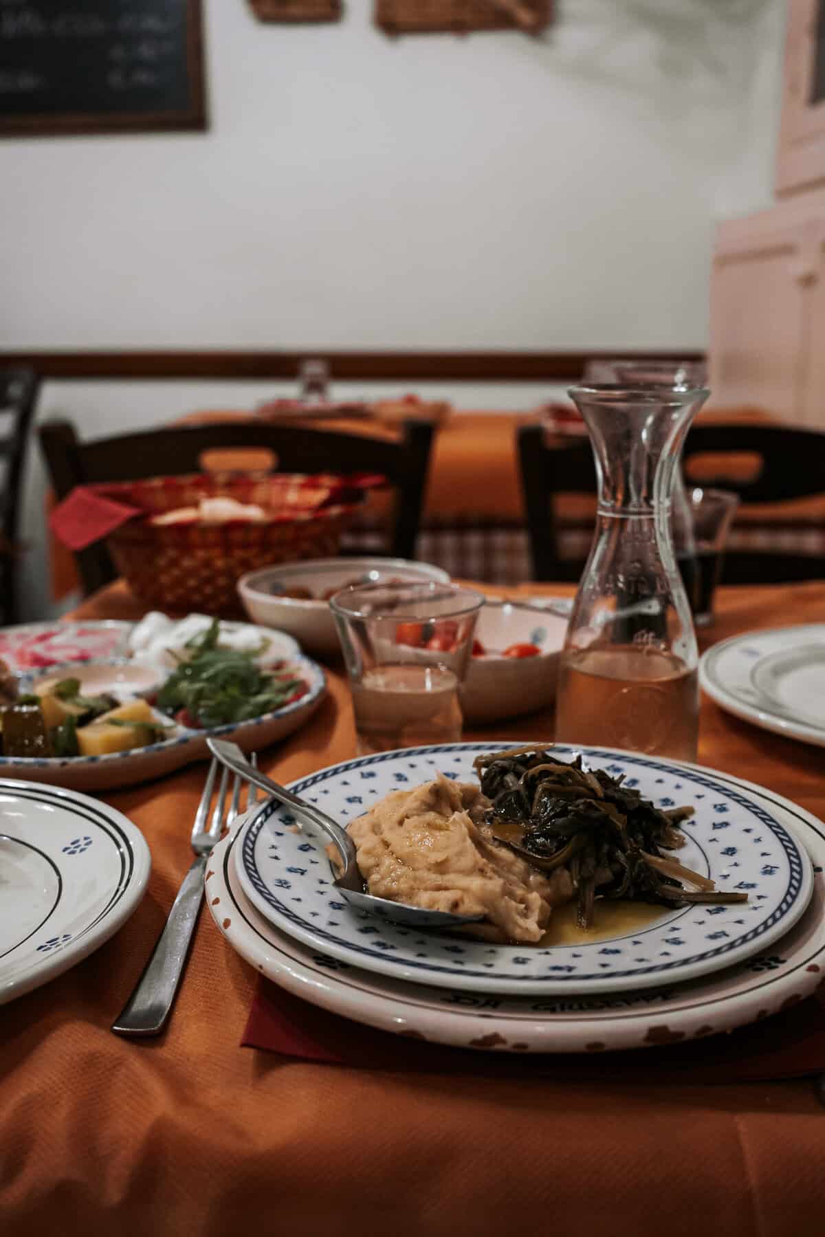 A cozy restaurant table set with a plate of traditional fava bean purée topped with sautéed chicory, surrounded by an assortment of dishes including fresh salad, bread, and a carafe of white wine. The warm ambiance reflects a homely, authentic dining experience.