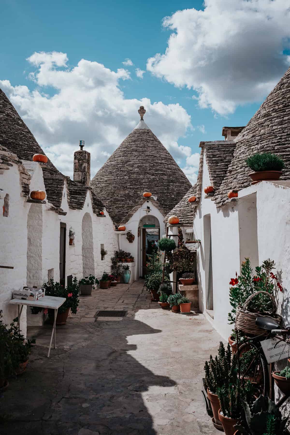 A charming alleyway in Alberobello, Italy, lined with traditional whitewashed trulli houses with conical stone roofs. The scene is full of potted plants and small pumpkins perched on rooftops, under a bright blue sky with fluffy clouds.