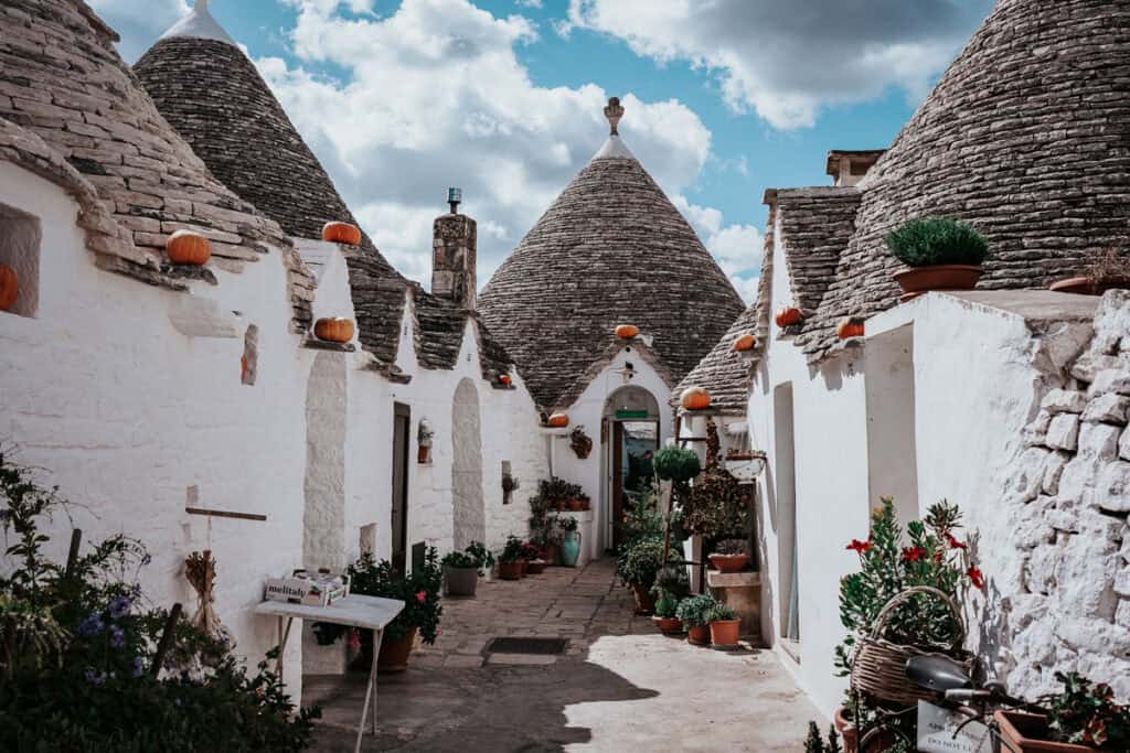 A frontal view of a narrow street in Alberobello showcasing rows of trulli houses with stone cone-shaped roofs and white walls. The walkway is decorated with terracotta pots of greenery and orange pumpkins, capturing a cozy and festive village vibe