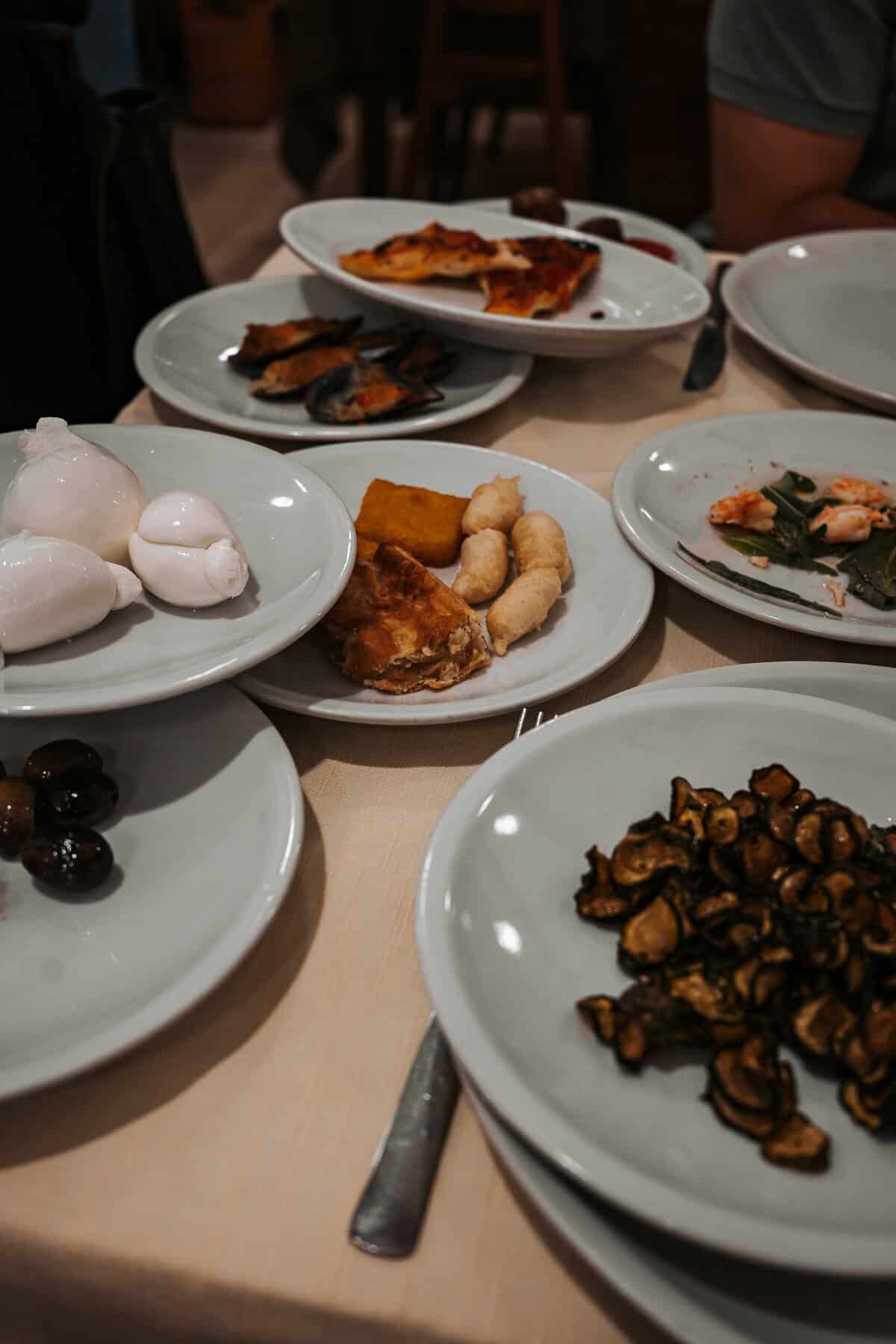 A spread of Italian antipasti on a restaurant table, including mozzarella balls, grilled zucchini, olives, fried dough, seafood, and slices of focaccia. The plates are arranged on a cream-colored tablecloth, showcasing a rustic and hearty meal.