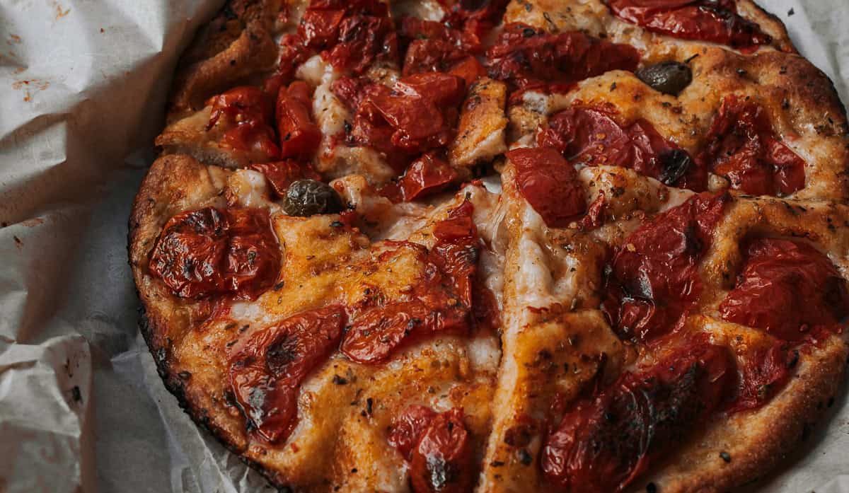 A rustic round focaccia topped with baked cherry tomatoes and green olives is sliced and laid out on parchment, set against stone pavement in an outdoor setting.
