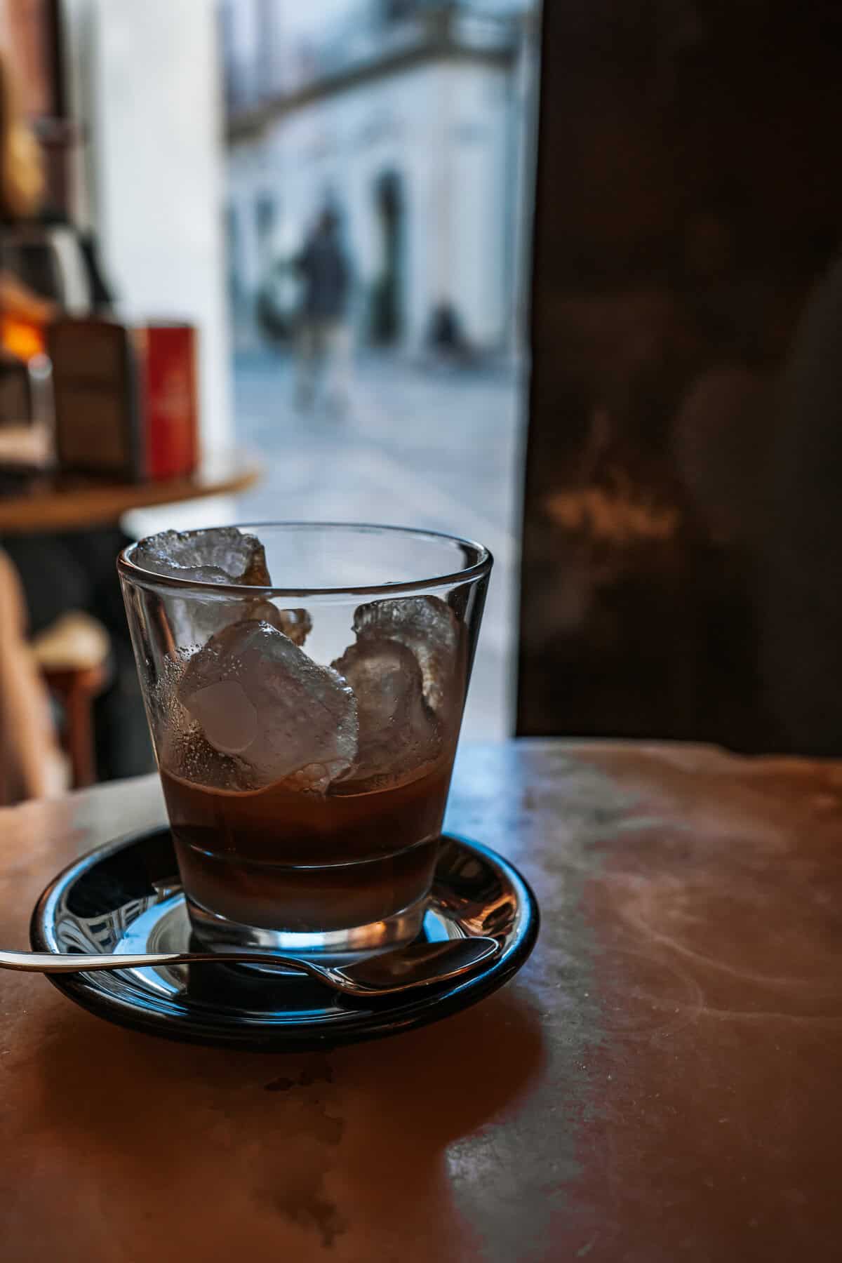 A glass of iced espresso sits on a black saucer with a spoon, catching the afternoon light in a cozy café with a blurred view of the street outside.