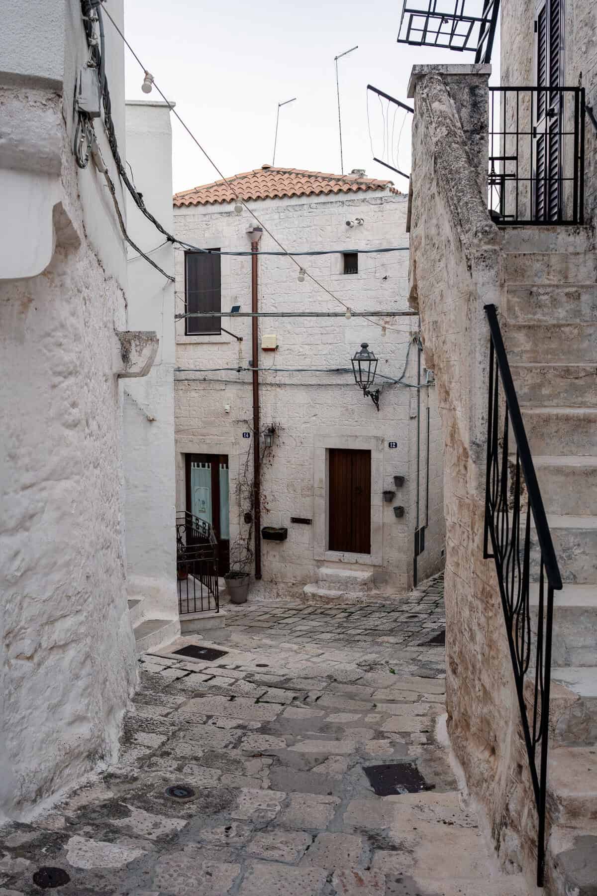 A quiet, narrow street in Ceglie Messapica, Puglia, flanked by stone buildings with weathered white façades and shuttered windows. String lights hang overhead, adding charm to the rustic alley and cobblestone path.