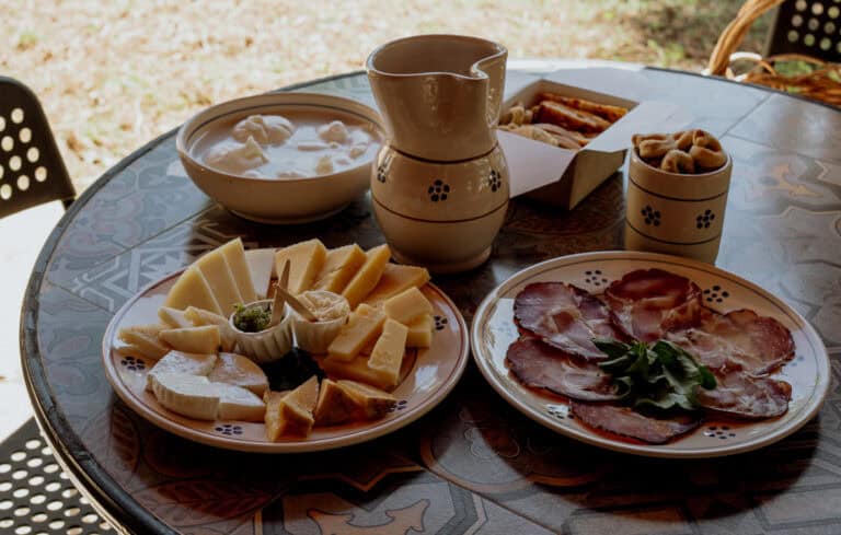 A rustic table set outdoors features a spread of local cheeses, cured meats, taralli, and a jug of milk, showcasing a traditional Italian antipasto under natural light.